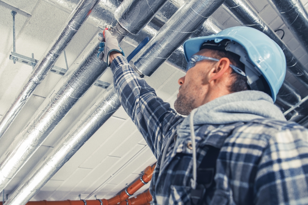 A worker inspects overhead pipes in a construction environment, ensuring safety compliance